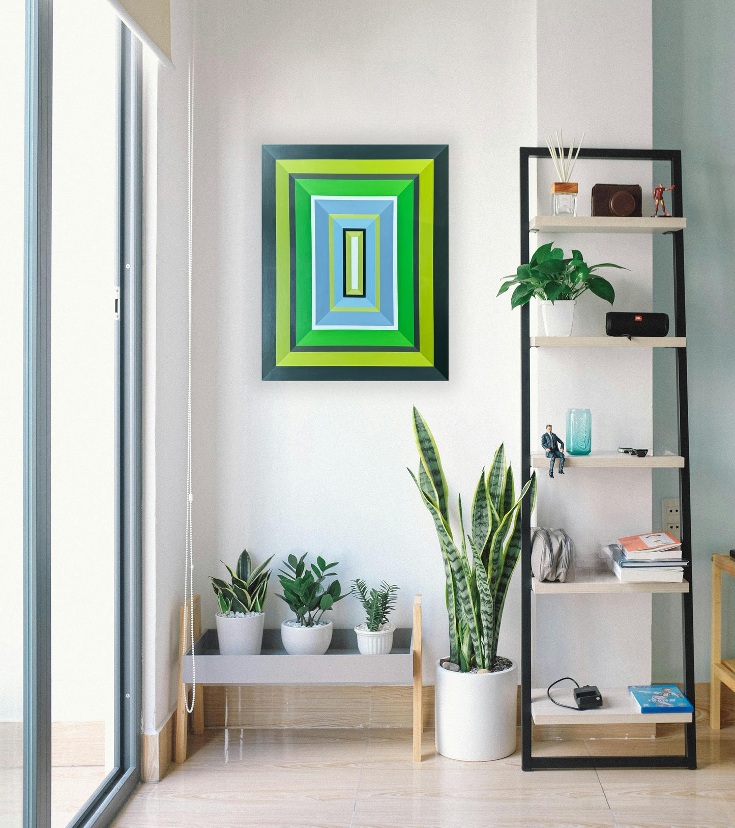 Hard edge abstract painting by Elena Alexander styled in a contemporary plant corner. The geometric wall art features concentric rectangles in flat colors - from deep charcoal through vibrant lime, emerald green, and serene blue, with a white-centered black frame. The painting's crisp lines and fresh color palette perfectly complement an urban jungle aesthetic, shown alongside snake plants, pothos, and other tropical houseplants. A modern minimalist piece demonstrating precise hard edge technique while harm