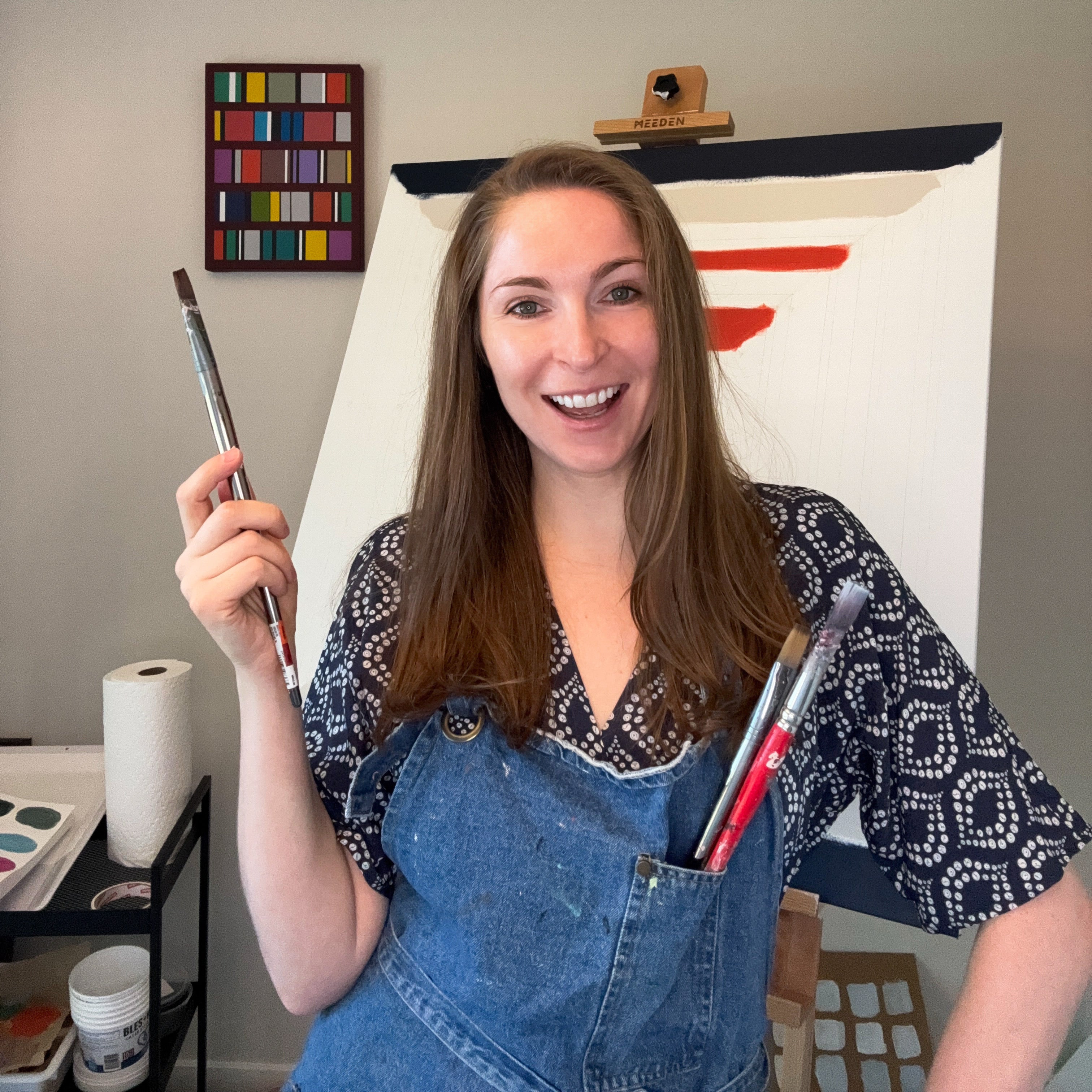 Artist Elena Alexander smiling in her studio, wearing denim apron and holding up a paintbrush to the camera.