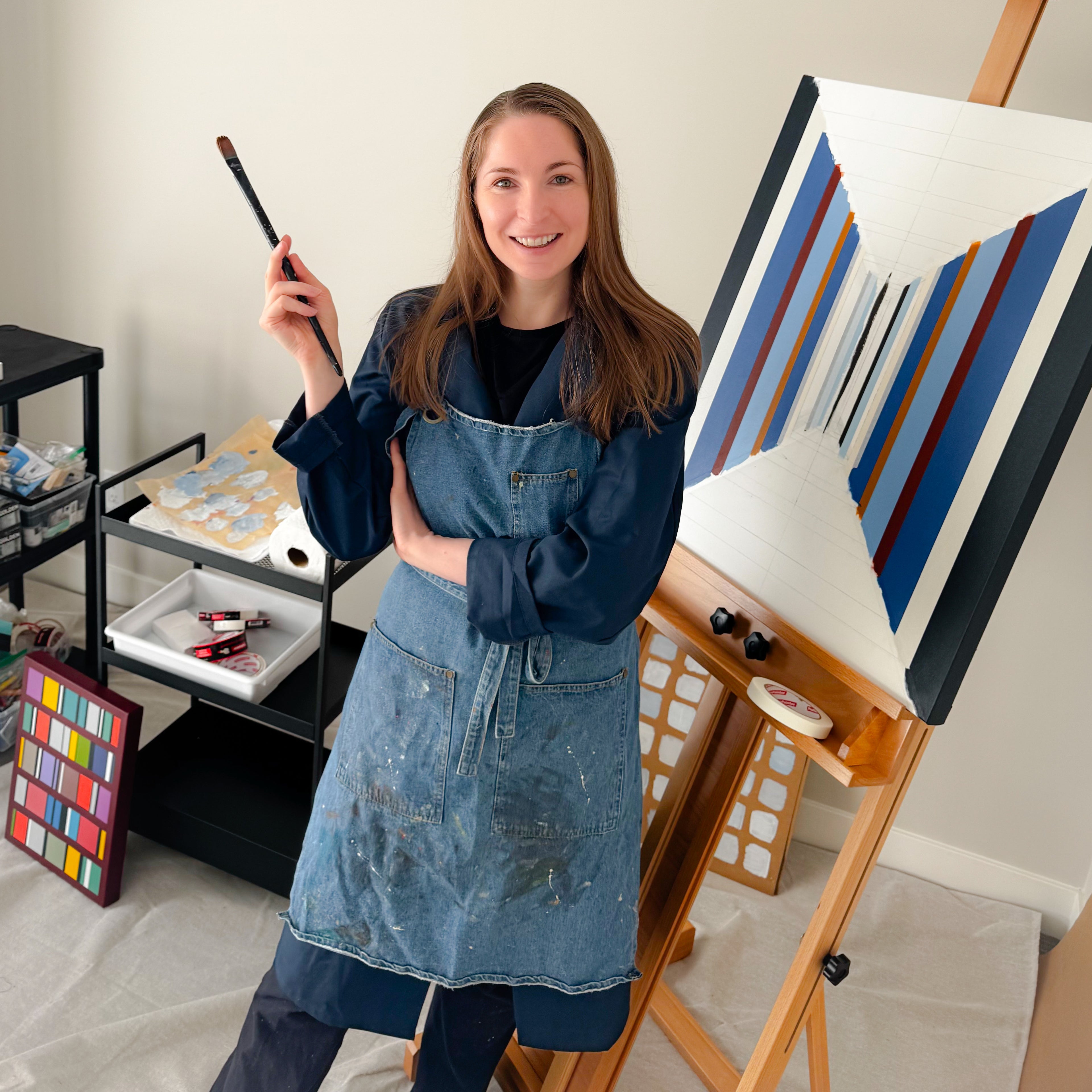 Elena Alexander in a denim apron holding a paintbrush next to an easel with a painting on it, in her art studio.