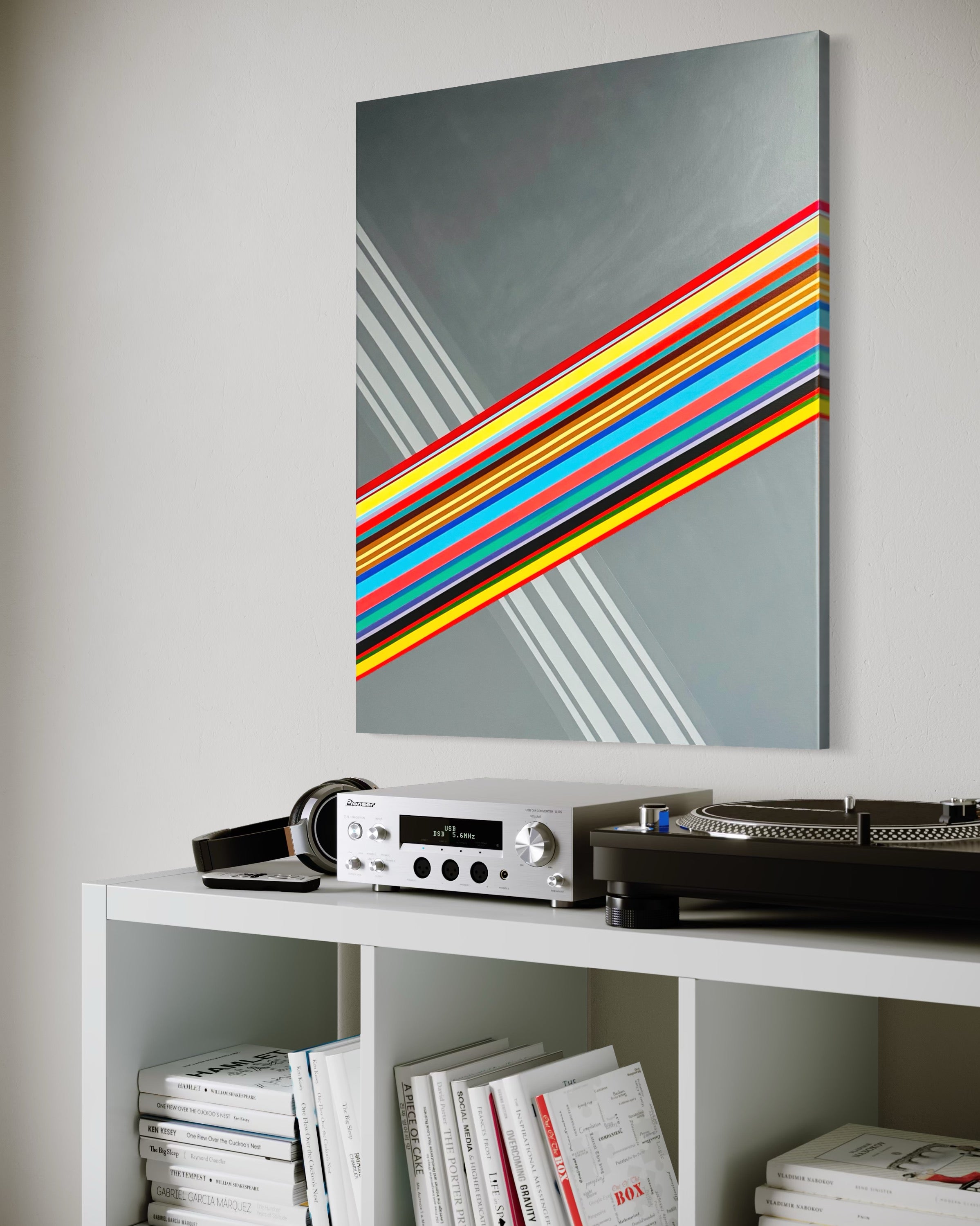 Colorful abstract art piece on a wall above a white shelf with electronic equipment and books.