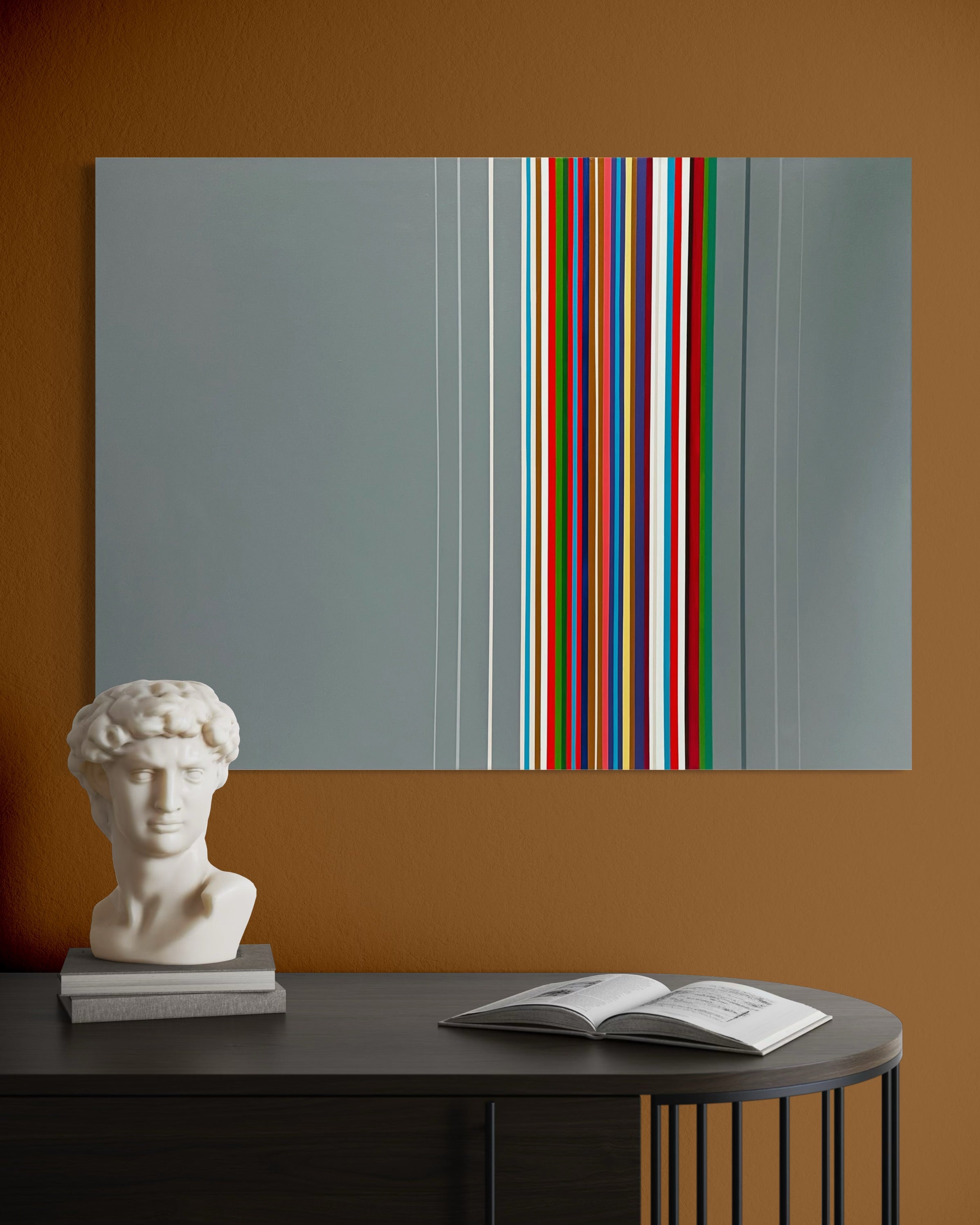 Abstract art piece with colorful stripes on a wall above a table with a book and statue.